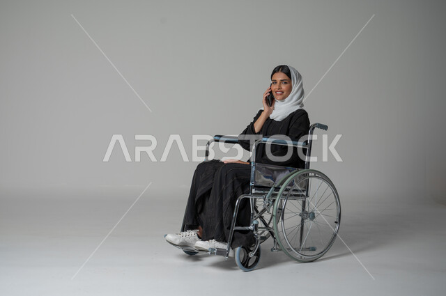 التواصل والاطمئنان على الأصدقاء، بورتريه مقرب لامرأة عربية سعودية خليجية من ذوي الاحتياجات الخاصة ترتدي العباءة السوداء والحجاب تجلس على كرسي متحرك تقوم بإجراء مكالمة صوتية عبر الجوال، الإجابة والرد على الاتصالات بإستخدام الهاتف المحمول، خلفية رمادية