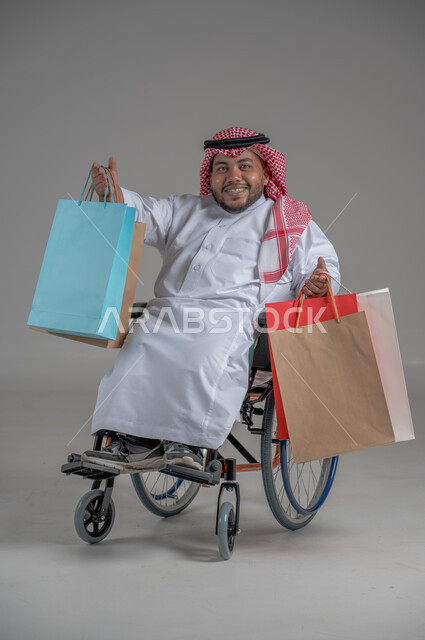 مفهوم التسوق والتبضع, ذوي الإحتياجات الخاصة والهمم, بورتريه لشاب عربي خليجي سعودي قزم مقعد يرتدي الثوب التقليدي والشماغ يجلس على كرسي متحرك ويحمل بيديه مجموعة من الاكياس الملونة, صورة الجسم بالطول الكامل، خلفية رمادية