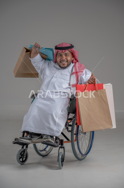 مفهوم التسوق والتبضع, ذوي الإحتياجات الخاصة والهمم, بورتريه لشاب عربي خليجي سعودي قزم مقعد يرتدي الثوب التقليدي والشماغ يجلس على كرسي متحرك ويحمل بيديه مجموعة من الاكياس الملونة, صورة الجسم بالطول الكامل، خلفية رمادية
