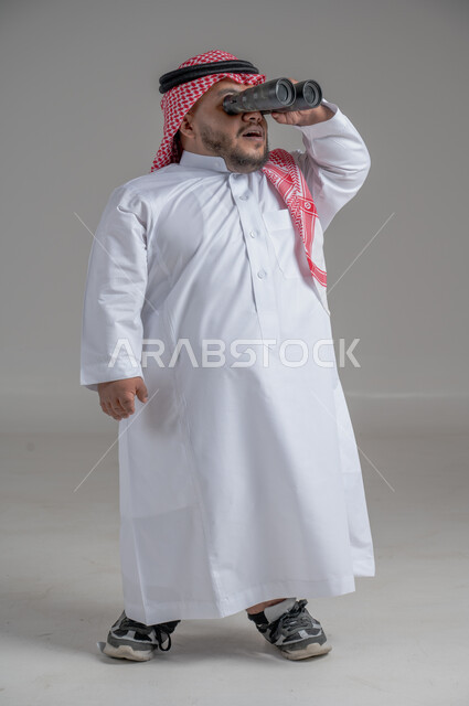 الشغف وحب الاستكشاف والمغامرة، البحث عن شيء ما باستخدام الناظور، مفهوم رؤية الأشياء البعيدة، بورتريه لرجل عربي خليجي سعودي قصير القامة يرتدي الثوب التقليدي والشماغ يقف باستقامة ينظر من خلال التلسكوب, حلم تحقيق الأهداف والطموحات المستقبلية, خلفية رمادية