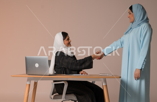 New partnership deals to expand the scope of work, accomplishing the required tasks and plans, portrait of two veiled Saudi Arabian Gulf women wearing the abaya shaking hands with gestures of pleasure for the success of the company's plan, cooperation and achieving high gains and profits, beige background
