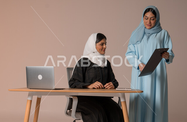 Cooperation between employees to achieve company goals, follow up and discuss business projects, portrait of two Saudi Arabian Gulf women completing work tasks and achieving the required goals, office jobs and professions, beige background