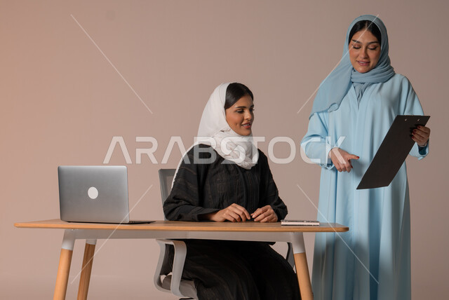 التعاون بين الموظفين لتحقيق أهداف الشركة، متابعة مشاريع الأعمال ومناقشتها، بورتريه لسيداتان عربيتان خليجيتان سعوديتان يقومان بإنجاز مهام العمل وتحقيق الأهداف المطلوبة، مهن ووظائف مكتبية، خلفية بيج