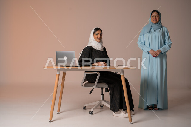 نمط الحياة وبيئة العمل السعودي، إمرأتان عربيتان خليجيتان سعوديتان محجبتان داخل مقر الشركة تنظران إلى الكاميرا بايماءات السعادة والسرور، العمل الجماعي وروح الفريق الواحد، الجسم بالطول الكامل، خلفية بيج