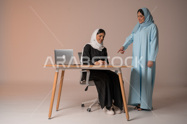 Teaching work methods and professional development, following up on employee task achievements, two veiled Saudi Arabian Gulf women developing plans and solving problems to achieve company goals, paying attention to and writing down notes, Saudi lifestyle and work environment, full length body, beige background
