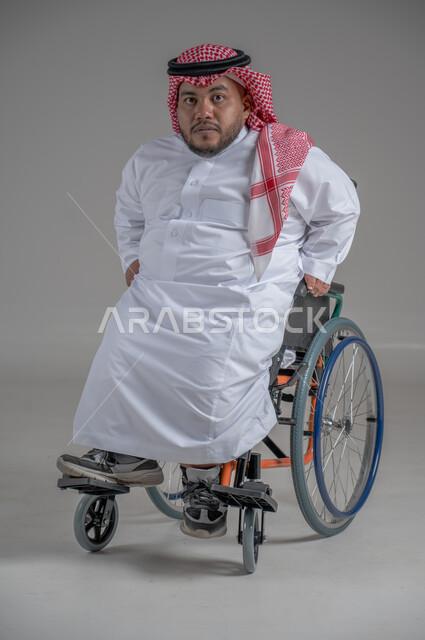 نمط حياة ذوي الهمم، استخدام كرسي المعاقين للمساعدة على الحركة والتنقل، بورتريه لشاب عربي خليجي سعودي قزم من ذوي الهمم يرتدي الثوب التقليدي والشماغ يجلس على الكرسي المتحرك ينظر الى الكاميرابايماءات السعادة والسرور، خلفية رمادية