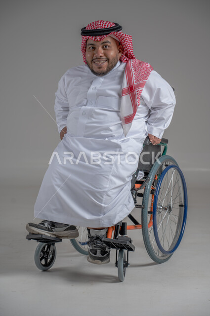 نمط حياة ذوي الهمم، استخدام كرسي المعاقين للمساعدة على الحركة والتنقل، بورتريه لشاب عربي خليجي سعودي قزم من ذوي الهمم يرتدي الثوب التقليدي والشماغ يجلس على الكرسي المتحرك ينظر الى الكاميرابايماءات السعادة والسرور، خلفية رمادية