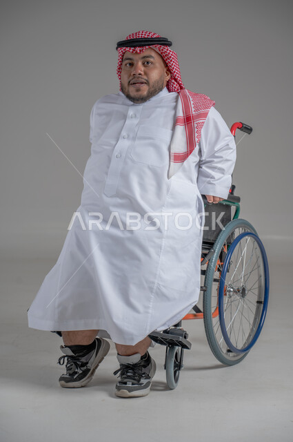 نمط حياة ذوي الهمم، استخدام كرسي المعاقين للمساعدة على الحركة والتنقل، بورتريه لشاب عربي خليجي سعودي قزم من ذوي الهمم يرتدي الثوب التقليدي والشماغ يجلس على الكرسي المتحرك ينظر الى الكاميرابايماءات السعادة والسرور، خلفية رمادية