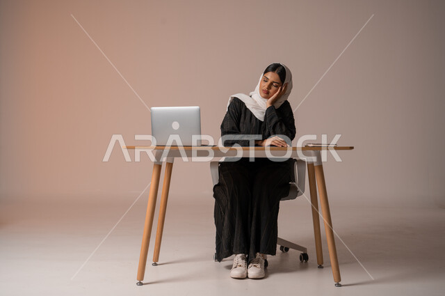 تعابير الكسل والخمول، الحاجة للنوم والراحة، بورتريه لإمرأه عربية خليجية سعودية محجبة ترتدي عباءة سوداء تجلس على الكرسي أمام مكتبها تضع يدها تحت رأسها بإيماءات تدل على النعاس والتعب، السهر لوقت متأخر، الجسم بالطول الكامل، خلفية بيج