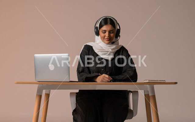 الرد على الاستعلامات والاستفسارات وخدمة العملاء، ايماءات التركيز بالعمل، بورتريه لامراة عربية خليجية سعودية محجبة ترتدي العباءة السوداء تجلس على الكرسي وتضع سماعة الهيدفون تمسك بيدها قلم وتدون الملاحظات المهمة، التواصل مع الأهل و الأصدقاء عن بعد، خلفية بيج