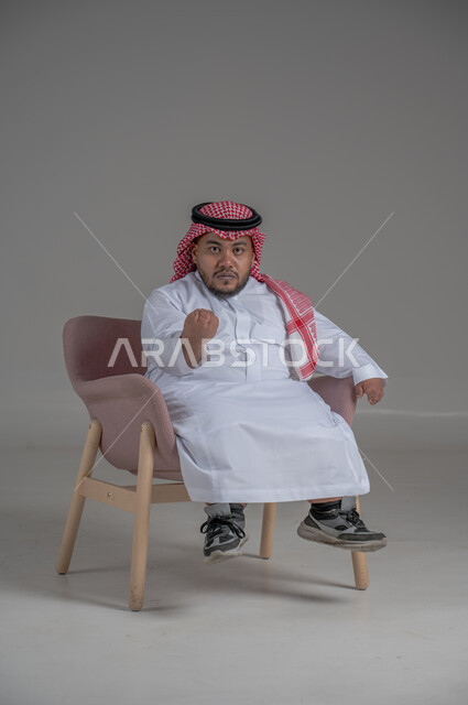 مفهوم الاناقة والاهتمام بالمظهر الخارجي، نمط حياة ذوي الهمم، بورتريه لشاب عربي خليجي سعودي يرتدي الثوب التقليدي والشماغ يجلس على كرسي مريح بايماءات السعادة والسرور، خلفية رمادية