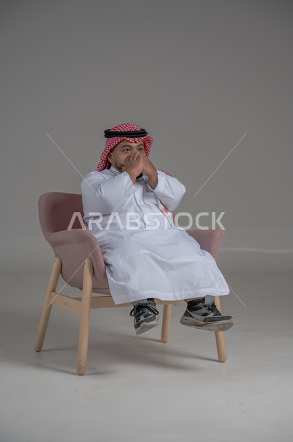 مفهوم الاناقة والاهتمام بالمظهر الخارجي، نمط حياة ذوي الهمم، بورتريه لشاب عربي خليجي سعودي يرتدي الثوب التقليدي والشماغ يجلس على كرسي مريح بايماءات السعادة والسرور، خلفية رمادية