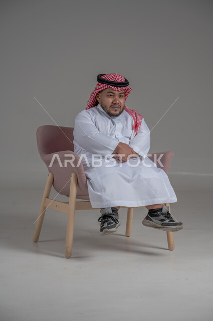 مفهوم الاناقة والاهتمام بالمظهر الخارجي، نمط حياة ذوي الهمم، بورتريه لشاب عربي خليجي سعودي يرتدي الثوب التقليدي والشماغ يجلس على كرسي مريح بايماءات السعادة والسرور، خلفية رمادية