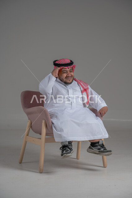 The concept of elegance and interest in appearance, lifestyle of people with disabilities, portrait of a young Saudi Arabian Gulf man wearing traditional thobe and shemagh sitting on a comfortable chair with gestures of happiness and pleasure, gray background