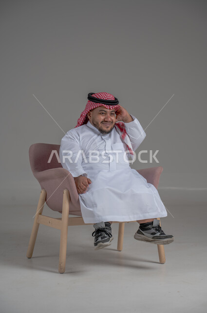 The concept of elegance and interest in appearance, lifestyle of people with disabilities, portrait of a young Saudi Arabian Gulf man wearing traditional thobe and shemagh sitting on a comfortable chair with gestures of happiness and pleasure, gray background