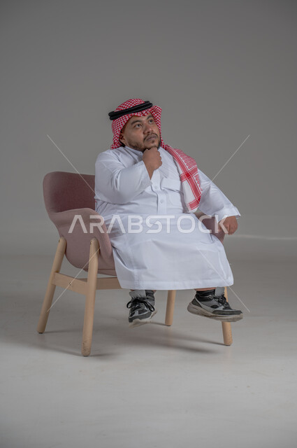 التخطيط للمستقبل والحياة، مفهوم التمعن والتركيز، حل المشكلات واتخاذ القرارات, ايماءات التأمل والتفكير والحيرة، نمط حياة ذوي الهمم،  بورتريه لشاب قزم قصير القامة عربي خليجي سعودي يرتدي الثوب التقليدي والشماغ يجلس على كرسي مريح يضع يده على ذقنه، خلفية رمادية