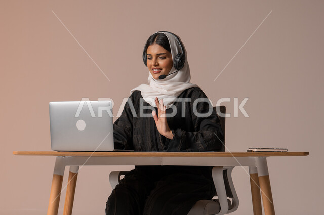مكالمات فيديو خلال اجتماع العمل, تمكين عمل المرأة في المجتمع السعودي,  بورتريه لإمرأة عربية خليجية سعودية محجبة ترتدي عباءة سوداء تضع سماعات الهيدفون تعمل في مجال خدمة العملاء, الرد على الاستعلامات والاستفسارات، استخدام التقنيات الحديثة والمتطورة، خلفية بيج