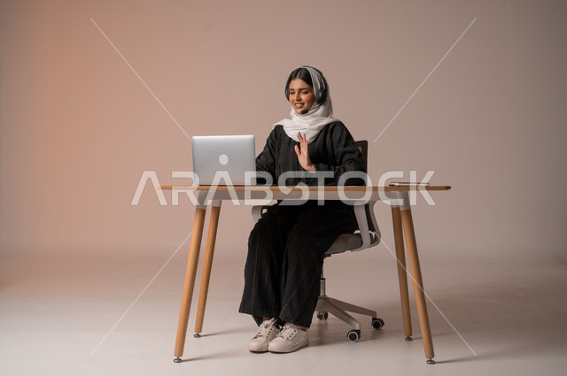 تمكين عمل المرأة في المجتمع السعودي,  بورتريه لإمرأة عربية خليجية سعودية محجبة ترتدي عباءة سوداء تضع سماعات الهيدفون تجري مكالمة فيديو خلال اجتماع العمل, الرد على الاستعلامات والاستفسارات، استخدام التقنيات الحديثة والمتطورة، الجسم بالطول الكامل، خلفية بيج