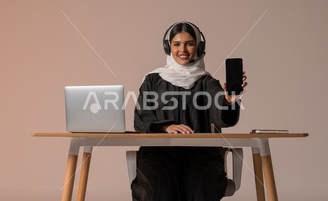 استخدام التقنيات الحديثة والمتطورة، الرد على الاستعلامات والاستفسارات وخدمة العملاء، بورتريه لامراة عربية خليجية سعودية محجبة ترتدي العباءة السوداء تجلس على الكرسي تمسك بيدها هاتف محمول بشاشة سوداء فارغة وتضع سماعة الهيدفون، التواصل مع الأهل و الأصدقاء عن بعد، خلفية بيج
