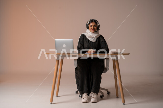 العمل في مجال خدمة العملاء والرد على الاسئلة والاستفسارات، الجلوس وراء المكتب وتنفيذ الأعمال المطلوبة, بورتريه لشابة عربية خليجية سعودية محجبة ترتدي عباءة سوداء تضع سماعات الهيدفون وتنظر للكاميرا بإيماءات السرور, الجسم بالطول الكامل, خلفية بيج