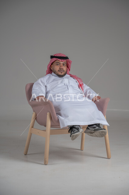 مفهوم الاناقة والاهتمام بالمظهر الخارجي، نمط حياة ذوي الهمم، بورتريه لشاب عربي خليجي سعودي يرتدي الثوب التقليدي والشماغ يجلس على كرسي مريح بايماءات السعادة والسرور، خلفية رمادية