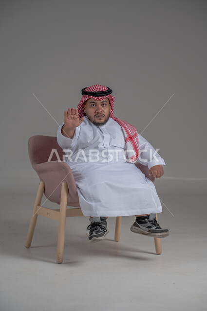 مفهوم الاناقة والاهتمام بالمظهر الخارجي، نمط حياة ذوي الهمم، بورتريه لشاب عربي خليجي سعودي يرتدي الثوب التقليدي والشماغ يجلس على كرسي مريح بايماءات السعادة والسرور، خلفية رمادية