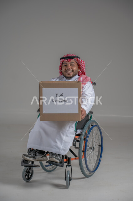 المساعدات والتبرعات الإنسانية، التشجيع على القيام بالأعمال الخيرية الإسلامية، الاحساس بالآخرين ومساعدتهم، بورتريه لشاب قزم قصير القامة عربي خليجي سعودي من ذوي الاحتياجات الخاصة يرتدي الثوب التقليدي يجلس على كرسي متحرك يحمل بيده صندوق تبرع،  خلفية رمادية