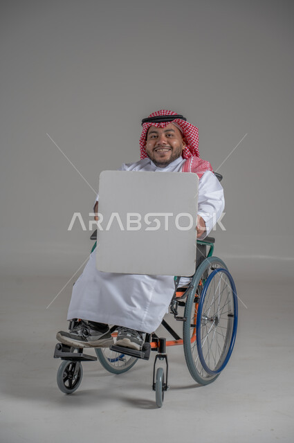 إعلانات وعروض تسويقية، موك اب مربع أبيض، استخدام لوحات توضيحية ووسائل تعليمية، بورتريه لشاب قزم قصير القامة عربي خليجي سعودي مقعد يرتدي الثوب التقليدي والشماغ يجلس على كرسي متحرك يحمل بيده لوحة بيضاء فارغة، خلفية رمادية
