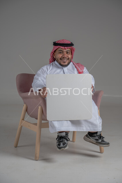 إعلانات وعروض تسويقية، موك اب مربع أبيض، استخدام لوحات توضيحية ووسائل تعليمية، بورتريه لشاب قزم قصير القامة عربي خليجي سعودي مقعد يرتدي الثوب التقليدي والشماغ يجلس على كرسي متحرك يحمل بيده لوحة بيضاء فارغة، خلفية رمادية