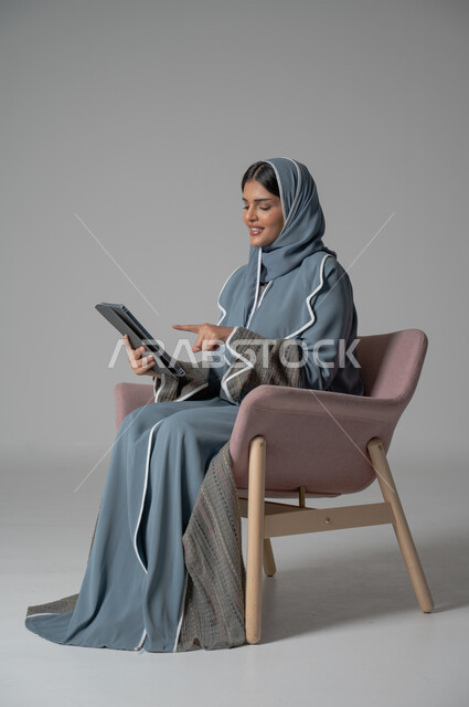 التسلية والترفيه وقضاء أوقات ممتعه, استخدام التكنولوجيا و الأجهزة التقنية الحديثة المتطورة, بورتريه لفتاة عربية خليجية سعودية محجبة ترتدي العباءة تجلس على كرسي مريح تحمل بيدها الموبايل وتشير اليه بايماءات السعادة والسرور, خلفية رمادية