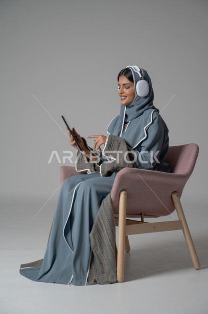 التسلية والترفيه وقضاء أوقات ممتعه, استخدام التكنولوجيا و الأجهزة التقنية الحديثة المتطورة, بورتريه لفتاة عربية خليجية سعودية محجبة ترتدي العباءة تجلس على كرسي مريح تضع سماعة الهيدفون وتحمل بيدها الموبايل وتشير اليه بايماءات السعادة والسرور, خلفية رمادية