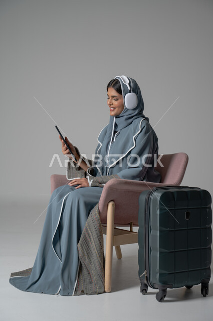 استخدام التكنولوجيا و الأجهزة التقنية الحديثة المتطورة, بورتريه لفتاة عربية خليجية سعودية محجبة ترتدي العباءة تجلس على كرسي مريح تحمل بيدها الموبايل وتضع سماعة الهيدفون بجانبها حقيبة السفر, ايماءات السعادة والسرور والاستمتاع بالتواصل عبر الجوال, خلفية رمادية