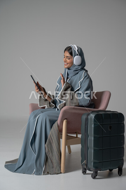 Gestures of happiness, pleasure, and enjoyment of communication via mobile phone, use of technology and advanced modern technical devices, portrait of a veiled Saudi Arabian Gulf girl wearing an abaya, sitting on a comfortable chair, holding the mobile phone in her hand and placing the headphone next to her travel bag, gray background
