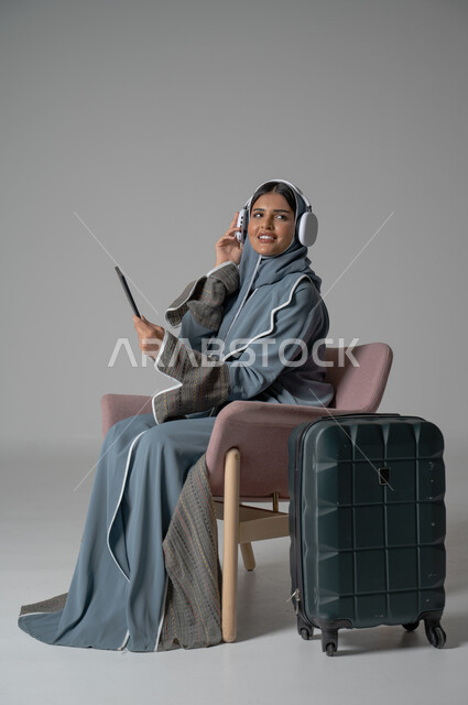 استخدام التكنولوجيا و الأجهزة التقنية الحديثة المتطورة, بورتريه لفتاة عربية خليجية سعودية محجبة ترتدي العباءة تجلس على كرسي مريح تحمل بيدها الموبايل وتضع سماعة الهيدفون بجانبها حقيبة السفر, ايماءات السعادة والسرور والاستمتاع بالتواصل عبر الجوال, خلفية رمادية