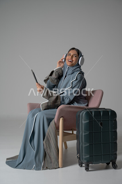Gestures of happiness, pleasure, and enjoyment of communication via mobile phone, use of technology and advanced modern technical devices, portrait of a veiled Saudi Arabian Gulf girl wearing an abaya, sitting on a comfortable chair, holding the mobile phone in her hand and placing the headphone next to her travel bag, gray background