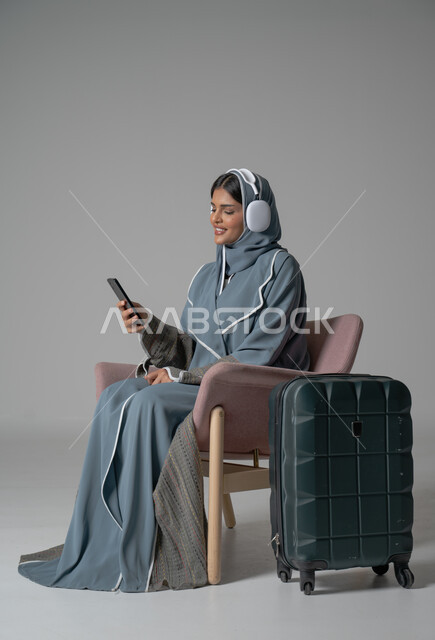 استخدام التكنولوجيا و الأجهزة التقنية الحديثة المتطورة, بورتريه لفتاة عربية خليجية سعودية محجبة ترتدي العباءة تجلس على كرسي مريح تحمل بيدها الموبايل وتضع سماعة الهيدفون بجانبها حقيبة السفر, ايماءات السعادة والسرور والاستمتاع بالتواصل عبر الجوال, خلفية رمادية