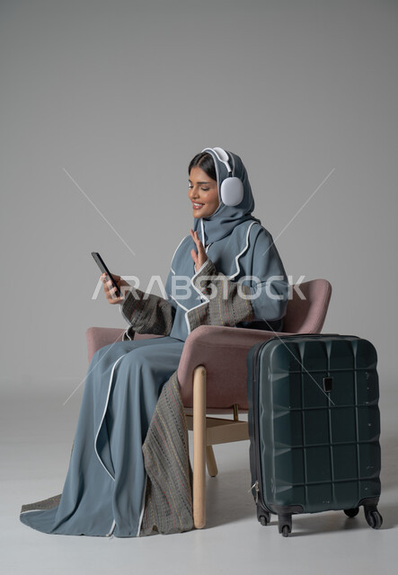   ايماءات السعادة والسرور والاستمتاع بالتواصل عبر الجوال, استخدام التكنولوجيا و الأجهزة التقنية الحديثة المتطورة, بورتريه لفتاة عربية خليجية سعودية محجبة ترتدي العباءة تجلس على كرسي مريح تحمل بيدها الموبايل وتضع سماعة الهيدفون بجانبها حقيبة السفر, خلفية رمادية
