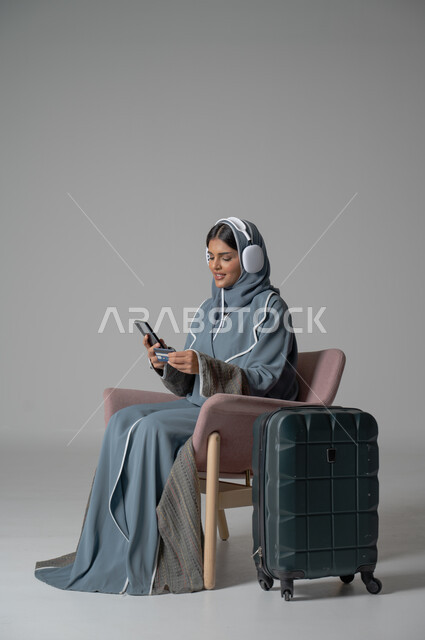الشراء والدفع عن طريق بطاقة الإئتمان البنكية, استخدام التكنولوجيا و الأجهزة التقنية الحديثة المتطورة, بورتريه لفتاة عربية خليجية سعودية محجبة ترتدي العباءة تجلس على كرسي مريح تحمل بيدها الموبايل وتضع سماعة الهيدفون بجانبها حقيبة السفر, خلفية رمادية