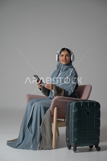 الشراء والدفع عن طريق بطاقة الإئتمان البنكية, استخدام التكنولوجيا و الأجهزة التقنية الحديثة المتطورة, بورتريه لفتاة عربية خليجية سعودية محجبة ترتدي العباءة تجلس على كرسي مريح تحمل بيدها الموبايل وتضع سماعة الهيدفون بجانبها حقيبة السفر, خلفية رمادية