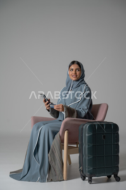 Purchase and payment via bank credit card, use of technology and advanced modern technical devices, portrait of a veiled Saudi Arabian Gulf girl wearing an abaya, sitting in a comfortable chair, holding a mobile phone in her hand next to her travel bag, gray background