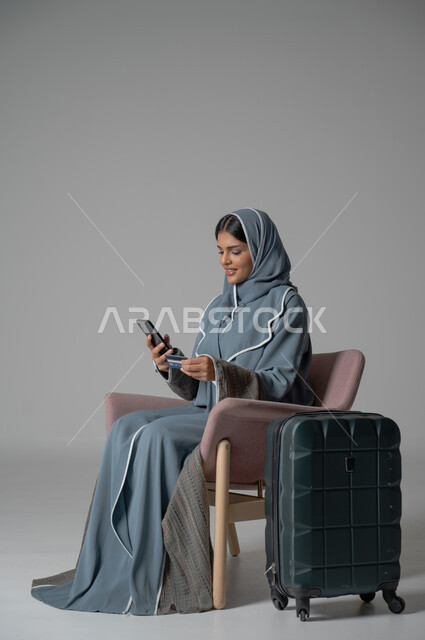 الشراء والدفع عن طريق بطاقة الإئتمان البنكية, استخدام التكنولوجيا و الأجهزة التقنية الحديثة المتطورة, بورتريه لفتاة عربية خليجية سعودية محجبة ترتدي العباءة تجلس على كرسي مريح تحمل بيدها الموبايل بجانبها حقيبة السفر, خلفية رمادية