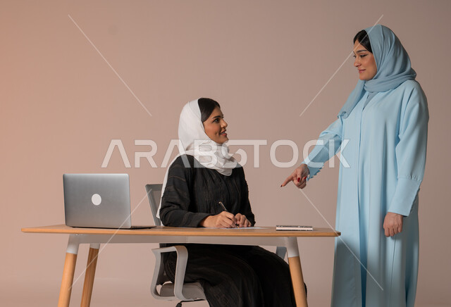 متابعة إنجاز مهام الموظفين، تعلم اساليب العمل والتطوير المهني، بورتريه لإمرأة عربية خليجية سعودية محجبة تشير باصبع السبابة الى ورقة تدوين الملاحظات, اعطاء التنبيهات الهامة للزملاء، مفهوم العمل التشاركي وروح الفريق الواحد، خلفية بيج