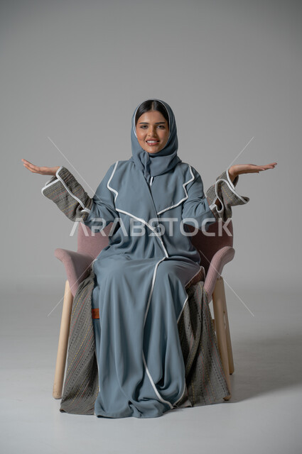 الاهتمام بالأناقة والمظهر الخارجي, بورتريه لامرأة عربية خليجية سعودية محجبة ترتدي العباءة تجلس على كرسي مريح تمد يديها جانبا بتعابير رفع عنصر ما, خلفية رمادية