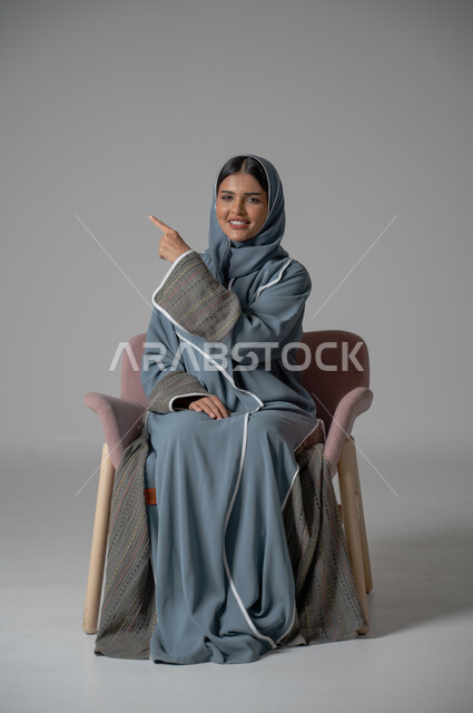 الاهتمام بالأناقة والمظهر الخارجي, بورتريه لامرأة عربية خليجية سعودية محجبة ترتدي العباءة تجلس على كرسي مريح تنظر الى الكاميرا تشير باصبع السبابة الى شيء ما, النظر الى الكاميرا بتعابير السرور, خلفية رمادية