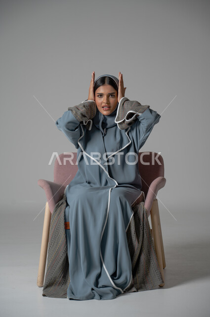 النظر الى الكاميرا بتعابير الحزن والخسارة, بورتريه لامرأة عربية خليجية سعودية محجبة ترتدي العباءة تجلس على كرسي مريح تضع يديها على رأسها بايماءات الدهشة والمفاجئة والانزعاج, خلفية رمادية