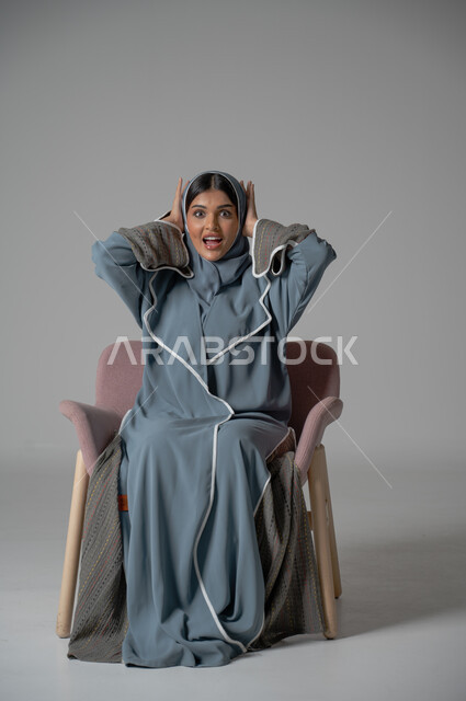النظر الى الكاميرا بتعابير الدهشة والفرح, بورتريه لامرأة عربية خليجية سعودية محجبة ترتدي العباءة تجلس على كرسي مريح تضع يديها على رأسها بايماءات الصدمة والمفاجئة لسماع خبر سار, خلفية رمادية