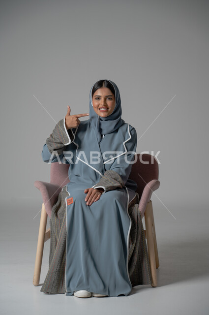 الاهتمام والعناية بالصحة الفموية، بورتريه لفتاة عربية خليجية سعودية محجبة ترتدي العباءة تجلس على كرسي مريح تنظر الى الكاميرا تشير بإصبعها على أسنانها، العناية والتجميل والتبييض، الزيارة الدورية إلى طبيب الأسنان، خلفية رمادية