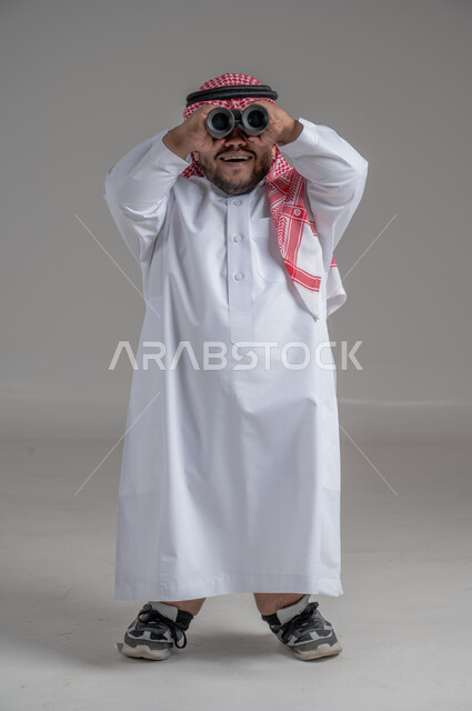 الشغف وحب الاستكشاف والمغامرة، البحث عن شيء ما باستخدام الناظور، مفهوم رؤية الأشياء البعيدة، بورتريه لرجل عربي خليجي سعودي قصير القامة يرتدي الثوب التقليدي والشماغ ينظر من خلال التلسكوب, حلم تحقيق الأهداف والطموحات المستقبلية, خلفية رمادية