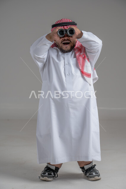 الشغف وحب الاستكشاف والمغامرة، البحث عن شيء ما باستخدام الناظور، مفهوم رؤية الأشياء البعيدة، بورتريه لرجل عربي خليجي سعودي قصير القامة يرتدي الثوب التقليدي والشماغ ينظر من خلال التلسكوب, حلم تحقيق الأهداف والطموحات المستقبلية, خلفية رمادية