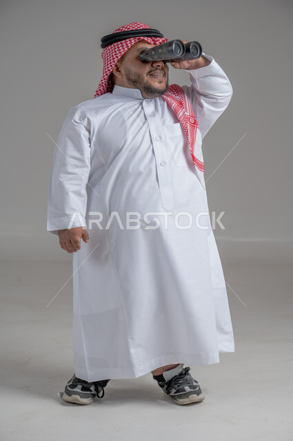 الشغف وحب الاستكشاف والمغامرة، البحث عن شيء ما باستخدام الناظور، مفهوم رؤية الأشياء البعيدة، بورتريه لرجل عربي خليجي سعودي قصير القامة يرتدي الثوب التقليدي والشماغ ينظر من خلال التلسكوب, حلم تحقيق الأهداف والطموحات المستقبلية, خلفية رمادية